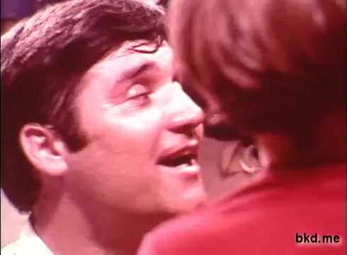 The still captures a moment from a 1970s Crest toothpaste commercial, featuring a man with a friendly expression and slightly tousled hair, possibly reacting to something or someone just off-camera. The background is softly blurred, directing focus on the man's face, which looks somewhat animated or enthusiastic. The color and grain reflect the vintage quality of a 16mm print, evoking a nostalgic feel typical of advertisements from that era.