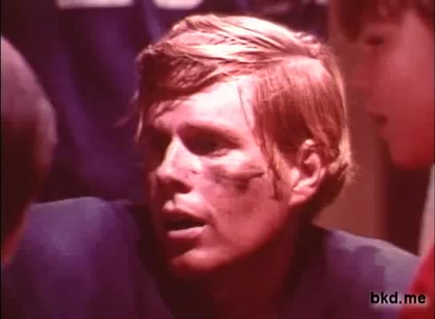 The still from the 1970s Crest toothpaste commercial features a young man with tousled hair and a somewhat rugged appearance. He has a dirty face with a smudge of dirt or grime, and a faint black mark, possibly makeup for effect. He appears to be engaged in conversation, with an attentive expression. The background shows hints of a team environment, suggesting themes of sports or camaraderie inherent in the commercial's context. The color tone reflects the vintage style of 16mm film.