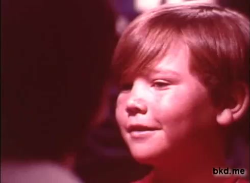The still depicts a young boy with light brown hair and a joyful expression, likely engaged in conversation. The background is softly blurred, characteristic of commercial aesthetics from the 1970s, emphasizing the boy's face. The color palette appears warm but slightly faded, typical of a 16mm print, giving it a nostalgic feel. The context suggests a friendly, cheerful environment, possibly aimed at highlighting the benefits of Crest toothpaste.