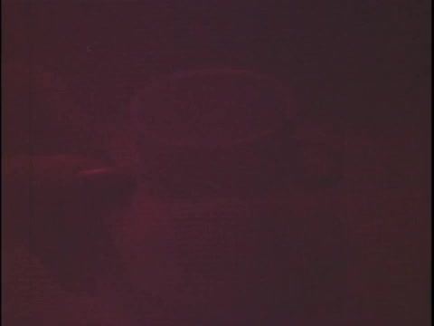 The still depicts a coffee cup situated in a coffee filter ring, with a dim, reddish hue dominating the image. The lighting creates a warm but muted ambiance, suggesting a focus on the coffee preparation process. The overall composition conveys a sense of warmth and coziness associated with coffee culture.