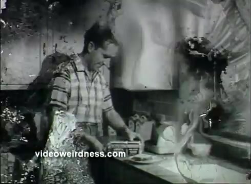 The still features a mid-20th-century kitchen scene, focusing on a man dressed in a plaid shirt as he prepares to wrap bread with Kaiser Aluminum foil. The image appears to have some wear, indicative of its age, and the kitchen background suggests a typical home setting of the 1950s. The commercial emphasizes the use of aluminum foil for freshness, aligning with the era's advertising style.