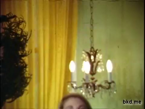 The still shows a vintage interior scene, possibly from a J.C. Penney commercial, featuring two individuals partially in frame. There’s a decorative chandelier with lit bulbs, and the background includes draped yellow curtains. The print quality suggests it is a 16mm film, capturing a nostalgic aesthetic typical of mid-20th century advertising.