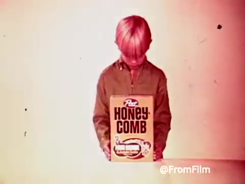 The still features a young boy with light hair, dressed in a green top, holding a box of Post Honey Comb cereal. The background has a retro, slightly faded look typical of 1960s film. This scene is part of a promotional segment for Archie's Alpha Bits cereal, highlighting the fun and appeal of the products during that era.