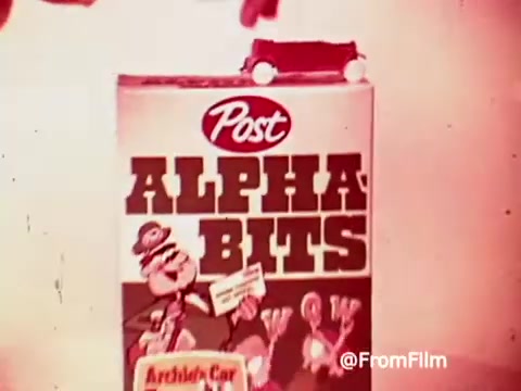 The still features a hand placing a small toy car on top of a cereal box labeled 'Post ALPHA BITS.' The box prominently showcases colorful graphics and characters associated with the cereal, tying in the promotional theme of the Archies' car. The image reflects the playful and vibrant marketing style typical of 1960s commercials.