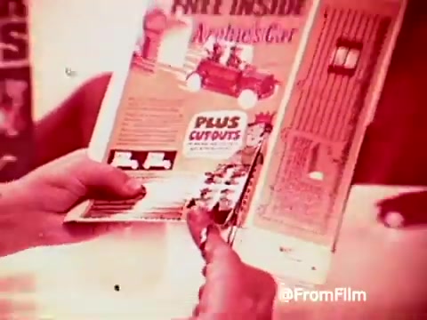 The still captures a hand holding a colorful promotional advertisement for Archies Alpha Bits cereal. The ad highlights a free toy, the 'Archies Car,' and features cutout instructions. The imagery has a vintage aesthetic, typical of 1960s commercials, with a warm color palette and playful graphics.