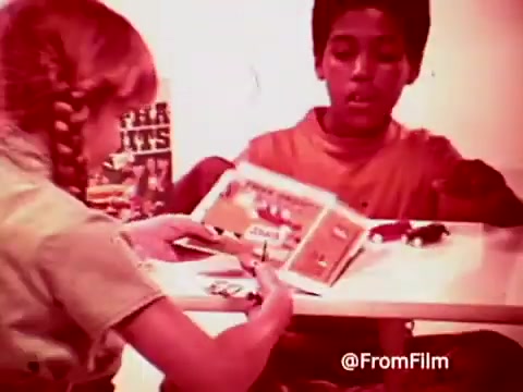 The still from the 1960s Archies Alpha Bits commercial features two children at a table. A girl with braids focuses on a cardboard playset, while a boy observes, sitting beside a box of Post Alpha Bits cereal. The scene captures the playful, vibrant energy typical of children's food advertising from that era.