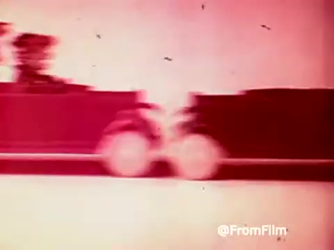 The still from the 1960s 'Archies Alpha Bits' commercial features a vibrant, stylized image of a cartoon vehicle, likely associated with the Archies. The car is depicted in motion, emphasizing a playful and energetic vibe, with a red and pink color palette that contributes to its nostalgic feel. The overall composition suggests a fun, child-friendly advertising approach for Post Alpha Bits cereal.