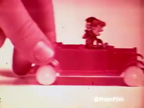 The still shows a playful scene from a 1960s Archies Alpha Bits commercial. A hand is positioned above a toy car featuring an animated character, likely from the Archie comics. The vibrant colors and whimsical design highlight the toy's appeal, capturing the essence of the cereal's promotion during that era. The overall vibe is cheerful and aimed at children, emphasizing fun and imagination.