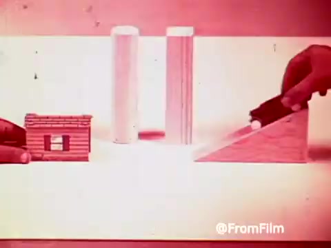 The still depicts a playful setup featuring hands manipulating a toy car on a ramp, alongside a small log cabin and two white cylinders. The scene captures the whimsical essence of a 1960s commercial promoting Archie’s Alpha Bits cereal, emphasizing fun and creativity. The color tone is soft and vintage, typical of 16mm prints from that era.