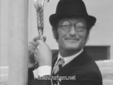 The 16mm still from 'Life and Breath' features a man in a bowler hat and round glasses, smiling as he holds onto a rope, suggesting a lighthearted or whimsical moment. The focus on his expression conveys a sense of joy or amusement within the context of the film. The black-and-white aesthetic enhances the retro feel of the scene.