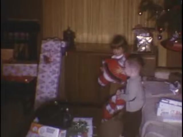 The still captures a moment from home movies at the Sisney house, featuring two young children amid a festive setting. They are surrounded by wrapped gifts and holiday decorations, suggesting a joyful occasion, likely Christmas. One child holds a toy or decoration, while the other appears to be excitedly approaching. The warm colors and cozy environment evoke a sense of nostalgia and family togetherness.