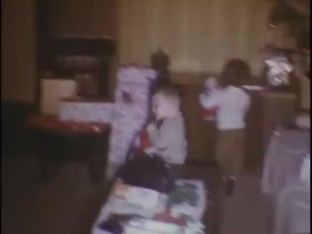 The still depicts a cozy interior scene, likely during a holiday gathering. Two young children are visible—one boy in a gray sweater and one girl in a white top—surrounded by colorful wrapped gifts. The atmosphere appears festive, suggesting a joyful occasion, possibly a Christmas celebration. The room features warm lighting and homey decor, evoking a sense of nostalgia.