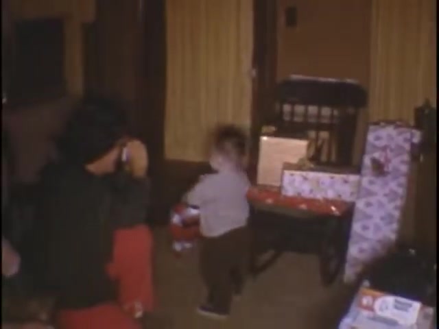 The still captures a nostalgic moment from a home movie at the Sisney house. A child, appearing to be around one or two years old, is seen playing with wrapped presents in a cozy living room setting. An adult, likely a family member, sits nearby, capturing the scene, possibly with a camera. The room features warm colors and homey décor, enhancing the intimate atmosphere of a family gathering.