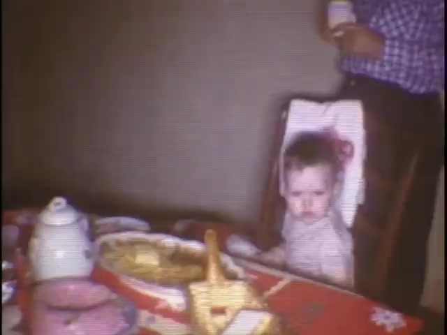 The still from 'home movies at the Sisney house' features a young child sitting in a high chair at a dining table. The table is adorned with a patterned tablecloth and contains various food items, including a large dish and a teapot. In the background, an adult can be seen holding a cup. The overall atmosphere appears casual and familial, capturing a moment of everyday life.