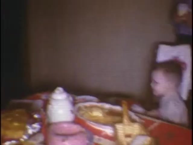The still captures a cozy scene at the Sisney house, featuring a young child sitting at a dining table. The table is filled with various food items, including pastries and a white container, suggesting a family gathering or meal. The background is dimly lit, emphasizing the warmth of the moment.