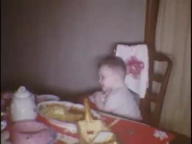 The still features a young child sitting at a dining table, possibly during a meal. The child, with short hair, appears to be in a high chair, with hands clasped together, suggesting a moment of anticipation or prayer. The table is adorned with a colorful tablecloth and various dishes, including bread. The warm, nostalgic tones convey a sense of family life and intimacy.