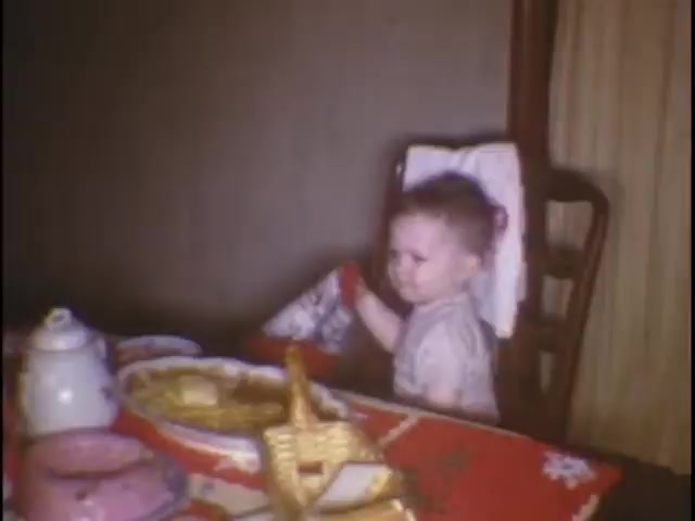The still captures a young child sitting at a dining table, surrounded by food items including a plate and a teapot. The child, wearing light clothing, appears to be playfully holding a napkin or cloth, possibly engaged in some playful activity. The setting suggests a cozy, domestic atmosphere typical of family home movies, likely during a meal or gathering at the Sisney home.