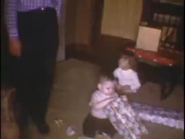The still captures a warm moment in a living room setting, where a young child is seated on the floor, focused on unwrapping a gift, while another child watches nearby. An adult, partially visible, stands beside them, indicating a family gathering. The atmosphere suggests a casual, joyful event. The decor hints at a cozy, lived-in space, enhancing the nostalgic feel of home movies.