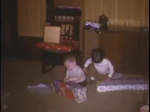 The still captures two young children on a carpeted floor, surrounded by colorful gift-wrapping. One child, a light-haired boy, is focused on a small present, while the other, wearing a white shirt, appears to be inspecting another gift nearby. The cozy interior features a wooden rocking chair and a cabinet, evoking a nostalgic home setting. The scene conveys a sense of childhood wonder and joy during a festive occasion.