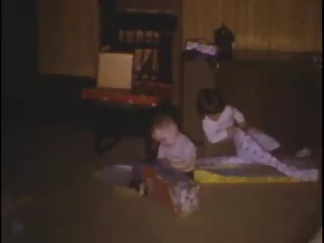 The still shows two young children playing on a carpeted floor in a cozy indoor setting. One child, a baby, is surrounded by colorful wrapped gifts, while the other, a slightly older child, is engaged in unwrapping a present. The room features warm lighting and simple furnishings, contributing to a nostalgic home movie atmosphere.