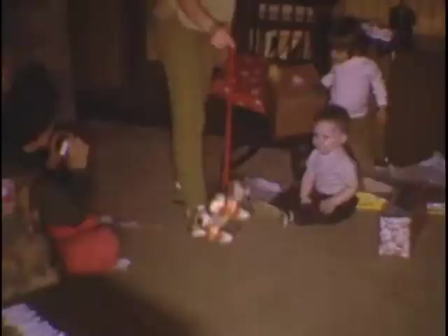 The still is a snapshot from 'home movies at the Sisney house,' capturing a cozy indoor scene. In a warm, domestic setting, a caregiver stands with a red toy cart, while two toddlers play on the carpet. One child sits cross-legged, looking at a stuffed toy, while another stands nearby, seemingly engaged. Toys and gifts are scattered around, suggesting a festive atmosphere, likely for a family gathering or celebration.