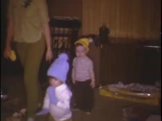 The still features two young children in a cozy indoor setting. One child is wearing a blue hat and gloves, while the other has a yellow hat. They appear to be playing or walking around, with an adult (presumably a caregiver) nearby. The room has a warm, nostalgic atmosphere, typical of home movies, with some toys and household items visible in the background.
