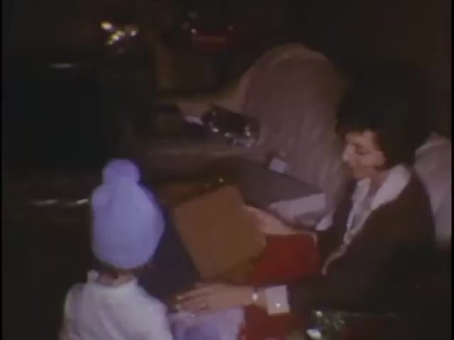 The still captures a cozy moment in the Sisney house, showing a woman seated and engaging with a young child. The child, wearing a light blue hat, appears to be receiving or looking at a box or gift. The setting is warm and familial, with soft lighting and a casual home environment, evoking a sense of nostalgia and intimate family connection.