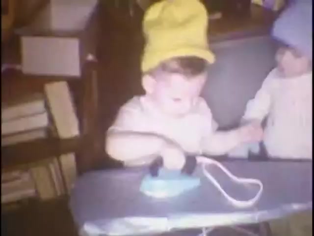 The still features a young child wearing a bright yellow hat, playfully pretending to use a blue toy iron on an ironing board. Another child, dressed in a light blue hat and sweater, appears to be engaging with the first child, creating a charming and playful scene. In the background, there are bookshelves, adding a cozy home atmosphere. This captures a moment from home movies, likely meant for reminiscing and sharing with family, particularly Susan.