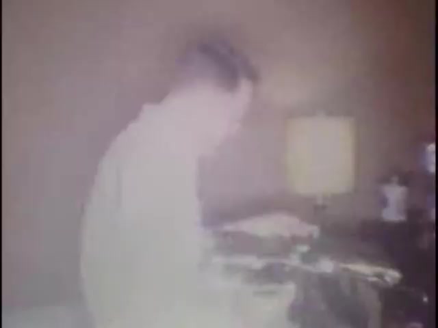 The still shows a man in a room, seemingly engaged with a radio or a similar device. The lighting is soft, and there is a lamp in the background, contributing to a homey atmosphere. Overall, it captures an intimate moment from home movies at the Sisney house, likely intended for Susan.