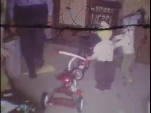 The still from 'home movies at the Sisney house' features a nostalgic scene with children playing indoors. Two young kids are engaged with a red tricycle, while an adult figure stands nearby. The overall ambiance suggests a warm, familial environment, capturing a moment of childhood joy and exploration. The image exhibits some wear typical of old film, adding to its vintage charm.