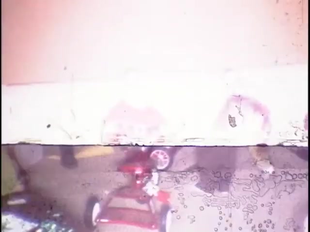 The still from 'home movies at the Sisney house' captures a nostalgic moment, likely featuring children playing with a small red toy car. The image shows signs of aging, with a washed-out color palette and visible scratches or distortion, enhancing its vintage feel. The focus is on a playful atmosphere, reflecting everyday family life.