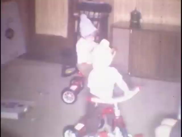 The still shows two young children riding small tricycles indoors. They are wearing knitted hats and appear to be having fun. The room features wooden paneling and some furniture in the background, creating a cozy home environment. This moment captures a playful, nostalgic scene from home movies intended for Susan.