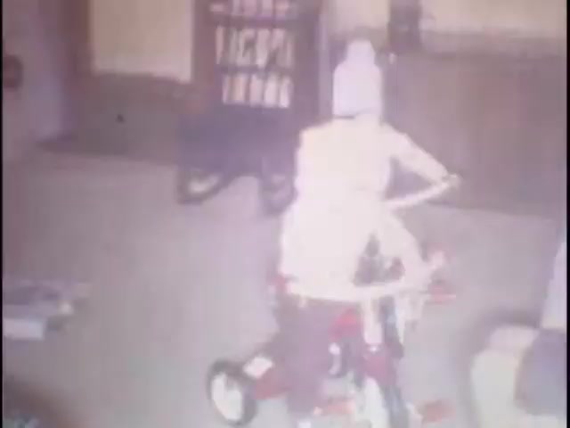The still features a child riding a small red tricycle in a cozy indoor setting. The child is wearing a hat, and the room has a nostalgic feel with bookshelves and soft lighting, suggesting a warm home environment. Various household items are scattered around, contributing to the casual atmosphere of home movies.