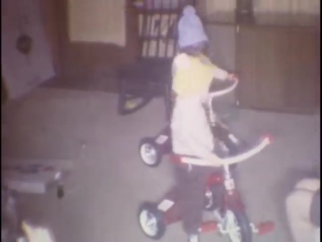The still captures a young child riding a red tricycle indoors. The child is wearing a light blue hat and a yellow shirt, with a white apron over them. The setting appears cozy, featuring a wooden railing in the background and a few scattered toys, evoking a nostalgic family moment.