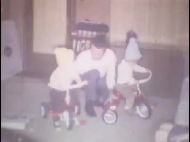 The still features two young children on red tricycles in a home setting, accompanied by an adult male. The children are wearing colorful winter hats, and the scene exudes a warm, cheerful atmosphere, likely capturing a playful moment in their home for Susan's memories. The background includes simple furnishings, contributing to the intimate, domestic feel of the moment.