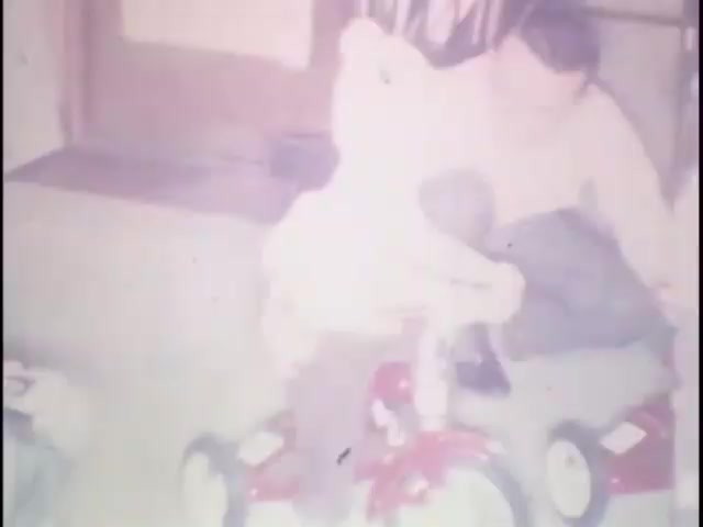 The still captures a moment from home movies at the Sisney house, featuring a young child riding a red tricycle. An adult, possibly a parent, is seated nearby, providing support or encouragement. The scene conveys a warm, nostalgic atmosphere, likely filled with family interactions and childhood joy. The lighting is soft, typical of vintage film, adding to the sentimental feel.