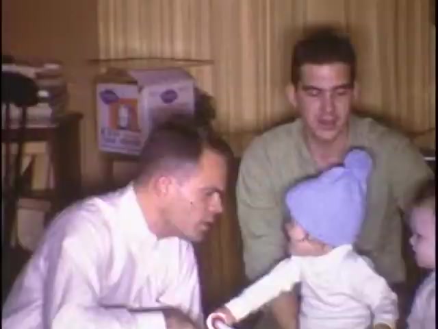 The still from 'Home Movies at the Sisney House' captures a warm family moment. Two adults are seen engaging with a toddler, who is wearing a blue knitted hat. One adult appears to be talking or playing with the child, while the other watches attentively. The background features boxes and curtains, providing a cozy domestic setting that highlights familial interaction.