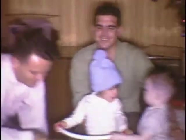 The still shows a warm, nostalgic scene from home movies at the Sisney house. Two adults appear engaged with two young children, one wearing a blue hat, creating a playful atmosphere. The backdrop suggests a cozy, festive setting, likely during a family gathering or celebration. The interaction conveys a sense of affection and joy among family members.