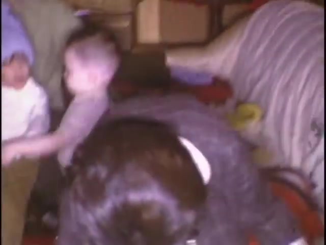 The still features a cozy indoor scene with two young children playing, one wearing a blue hat. A woman, partially visible, is seated nearby, engaged with the kids. The setting appears homey, with furniture and blankets in the background, suggesting a casual family gathering. The nostalgic feel of the home movies captures a candid moment of childhood and family connection.