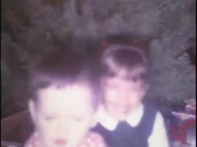 The still captures two young children in a cozy indoor setting, likely during a family gathering. One child, with short hair, appears out of focus and is seated in the foreground, while the other, a girl with a bow in her hair, smiles brightly in the background. A festive atmosphere is suggested by hints of greenery and wrapped gifts, evoking a nostalgic, homey vibe typical of family movie memories.