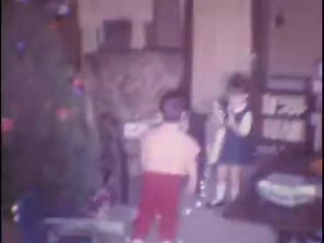 The still captures a nostalgic moment in a home setting, likely during a holiday season. A young boy, shirtless and wearing red pants, stands facing a girl in a blue dress with white trim, who appears to be holding something, possibly a toy. A decorated Christmas tree is visible in the foreground, setting a festive atmosphere. The background features a cozy living room with furniture and decor typical of the era, creating an intimate family scene.