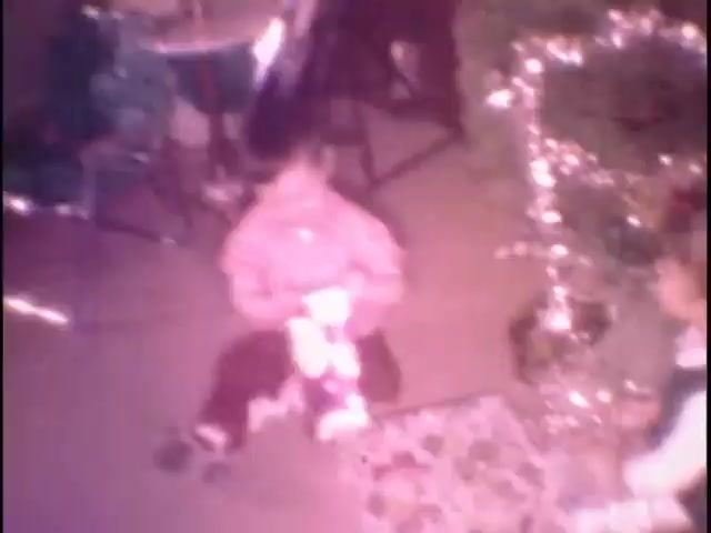 The still captures a nostalgic moment from home movies at the Sisney house, featuring a child sitting on the floor, holding a toy. The child is dressed in a pink outfit, while another individual, possibly an adult, is visible nearby. In the background, there are decorative lights and festive elements, suggesting a holiday setting. The warm, vintage tones evoke a sense of family and celebration.