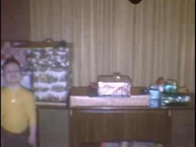 The still features a cozy indoor setting, likely during a festive occasion. In the foreground, a child stands smiling next to a low table adorned with several wrapped gifts. The background showcases patterned curtains, contributing to a nostalgic atmosphere typical of home movies. The scene captures a moment of joy and anticipation, presumably related to a celebration.