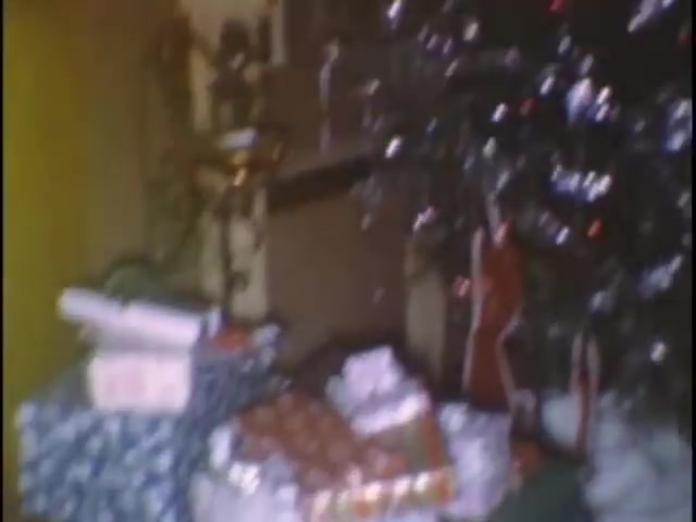 The still captures a cozy holiday scene at the Sisney house, featuring an assortment of wrapped gifts piled around a decorated Christmas tree. The warm, nostalgic atmosphere is enhanced by the presence of ornaments and tinsel on the tree, suggesting a festive celebration for Susan.