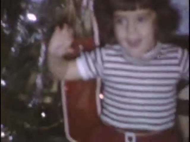 The still captures a young child, smiling and waving in front of a Christmas tree adorned with lights. They are dressed in a striped shirt and red shorts, exuding joy and excitement, likely enjoying a festive moment at the Sisney house during the holiday season. The warm, vintage quality of the 16mm film adds a nostalgic feel to the scene.