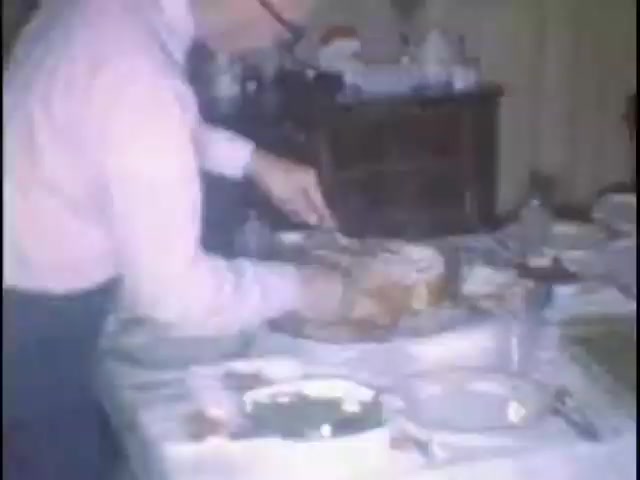The still captures a man in a pink shirt preparing to cut a cake or dessert on a dining table set with plates and utensils. The background features a dresser and other household items, suggesting a family gathering or celebration at the Sisney house, likely for Susan. The scene conveys a warm, domestic atmosphere.