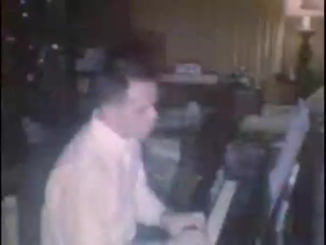 The still captures a man in a light-colored shirt playing a piano in a warmly lit room. The background features a cozy setting with furniture and decorations typical of a home environment, suggesting a personal atmosphere, possibly during a family gathering. The mood seems reflective and intimate, typical of home movies made for special occasions.