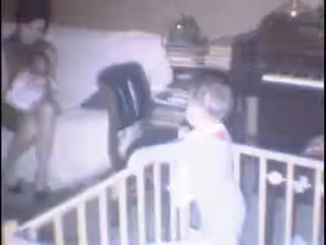 The still features a cozy indoor scene from home movies at the Sisney house. A woman sits on a couch, holding a baby, while a toddler stands in a playpen reaching out. The room is filled with a mix of furniture and books, conveying a warm, family-oriented atmosphere.