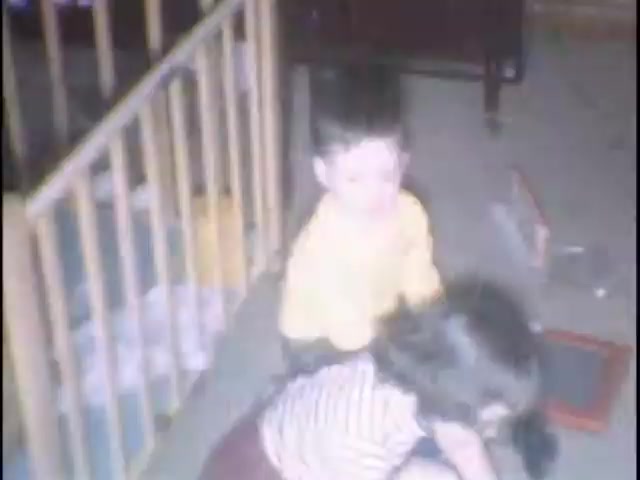 The still features two young children in a living room setting. The child in a yellow shirt appears to be playfully interacting with a girl who is wearing a striped shirt. They are on a carpeted floor, surrounded by toys and a playpen in the background, creating a warm, nostalgic atmosphere typical of home movies.