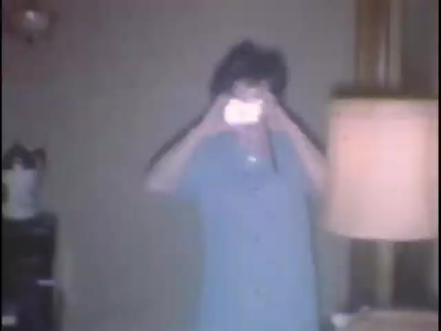 The still from 'home movies at the Sisney house' captures a woman holding a camera, likely taking a picture. She's dressed in a blue garment, standing in a living room setting with a lamp and what appears to be a decorative piece in the background. The overall atmosphere suggests a casual, intimate moment in a home environment.